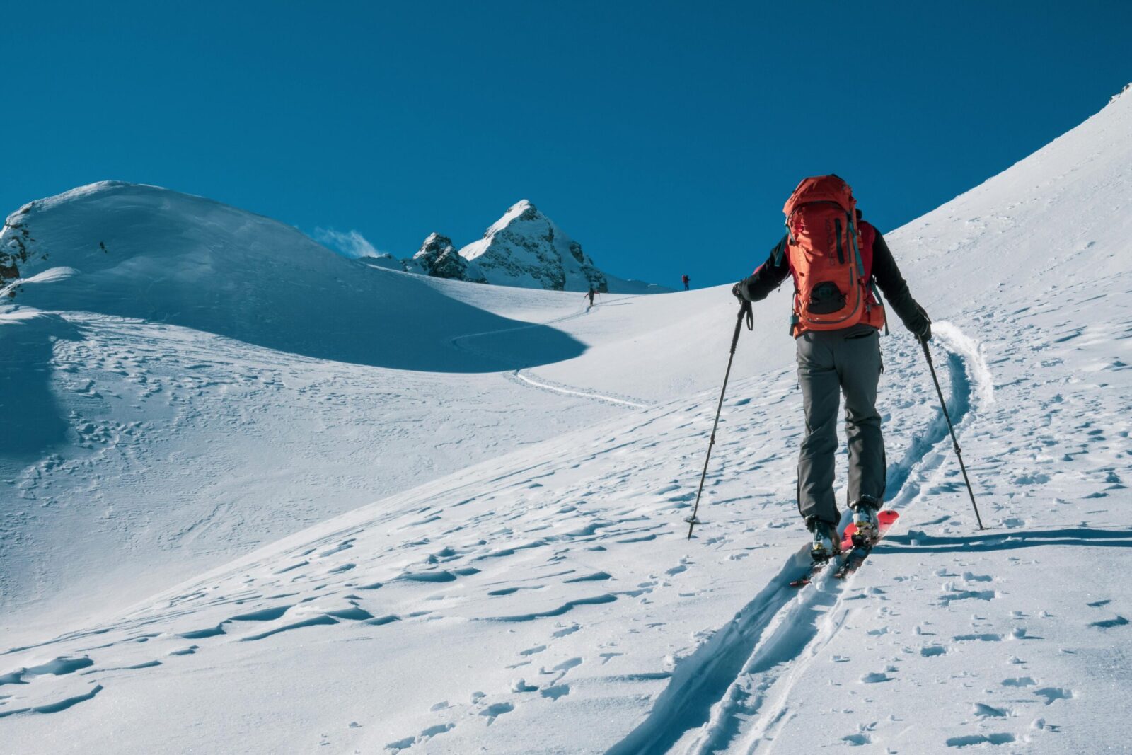 Person ski touring uphill in a snowy mountain landscape with clear blue sky.